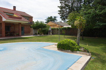 Fototapeta premium Pool covered with blue tarp in a green garden of a private house on a sunny day. End or beginning of season, fall or spring