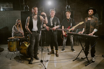 Group of rock male and female musicians playing in rock band posing for camera on stage in modern nightclub