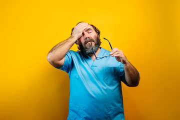 Doctor in blue coat on yellow background gesturing, makes tired gesture with hands on head.