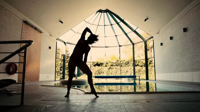 Silhouette of female acrobat doing handstand and leg pose next to indoor pool