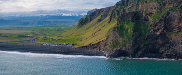 Dramatic aerial view of a steep, rocky cliff with green vegetation descending to a black sand beach, bordered by a turquoise ocean in Iceland.