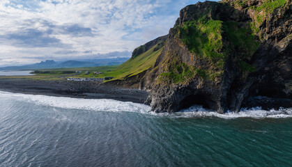 Obraz premium Stunning aerial view of Iceland's coastline showcasing a dramatic cliff with a cave, lush green vegetation, a black sand beach, and turquoise ocean waters.