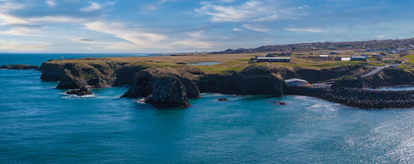 Stunning aerial view of Iceland's rugged coastline featuring cliffs, a small bay, and distant buildings under a clear blue sky with white clouds.