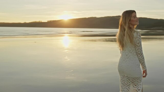 A woman in a white lace dress stands on a serene beach at sunset, gazing back with a gentle smile. The calm sea and distant islands create a tranquil and picturesque background.