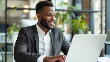 Fototapeta premium A happy Latin businessman in a suit sits at a desk in a modern office, smiling as he works on his laptop
