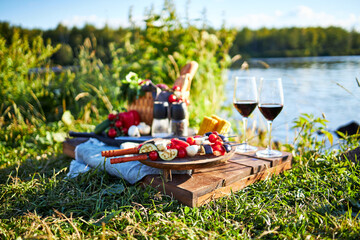 still life with vegetables, meat and glasses with red wine for picnic on a wooden tray on the river bank