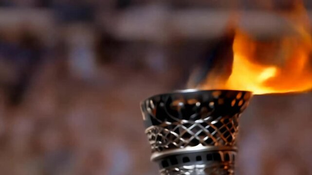 A close-up image of a gold Olympic torch with a burning flame in front of a blurred crowd