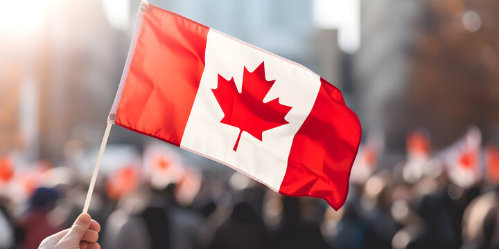 Close-up of a hand holding a small Canadian flag, blurred background of a city street, celebrating crowd