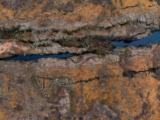 A stunning aerial perspective of a deep crack in the earth, filled with a narrow stream, surrounded by rocky, brown volcanic terrain in Iceland.