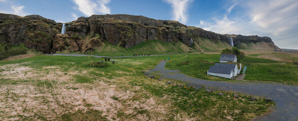 Stunning aerial view of a large cliff in Iceland with two waterfalls cascading down. Two white buildings and a gravel road are visible in the green and brown landscape.