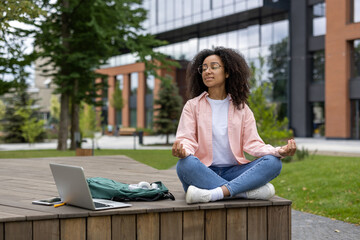 Young female student practicing meditation outdoors near university campus with laptop and backpack. Embracing mindfulness and relaxation amidst nature while taking a break from studies.
