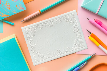 A colorful and creative workspace setup featuring a blank embossed card surrounded by various stationery items including pens, markers, a pencil, and decorative flowers, on a pastel background