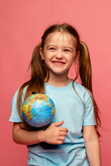 Happy smiling little schoolgirl girl in a blue t-shirt and denim shorts on a pink background with a globe in her hands. Copy space. Back to school concept. Earth Day
