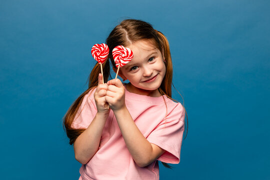 Happiness, childhood, kid and sweets. A happy little girl holding a heart shaped lollipop caramel on a stick, A festive candy in the hands of child in pink t-shirt on blue background. Valentines Day
