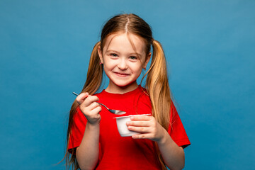 Cute little blonde hair girl in red t-shirt eating tasty yogurt on light blue background. Healthy...