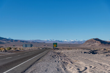 Road in southerly direction from San Pedro de Atacama, Antofagasta, Chile between sand and salt flats with in background snow covered the volcanic ridge with Argentina including Pular stratovolcano 