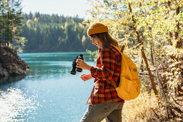 Young woman wildlife manager in red plaid shirt with yellow backpack with binoculars on lake shore...