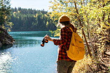 Young woman ecologist with a yellow backpack with binoculars on the rocky shore of a lake with blue...