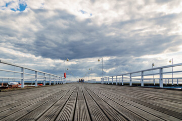 Fototapeta premium Seaside pier with beautiful stylish lanterns and people walking on it on a warm summer evening under cloudy sky