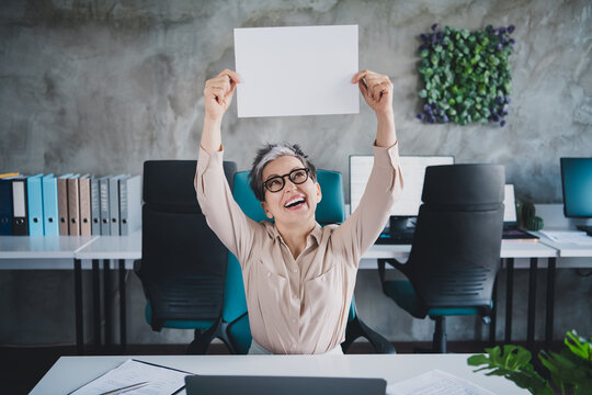 Photo of happy charming senior lady secretary wear formal shirt rising placard empty space indoors workshop workstation loft - Powered by Adobe