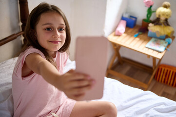 Beautiful little child girl 7 years old, smiling cutely while taking a self-portrait with smartphone on bed at home. The room is bright and cozy, capturing a candid and light-hearted moment.