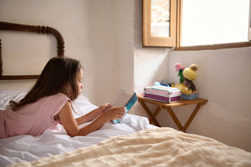 Relaxed girl in pink pajamas, reading book, lying on her belly on the bed in the morning. An open window on the background with falling sunbeams. The concept of World Book Day