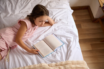 Top view calm relaxed girl in pink pajamas reading book or fairy tale, lying on the bed in the morning. The concept of a happy carefree childhood, education and erudition. Content for World Book Day