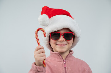 Portrait of a cute Caucasian girl in a Santa Claus hat and sunglasses eating a lollipop on a white background. 