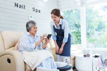 Asian female foot spa shop owner smiling while helping elderly Asian woman with smartphone in cozy nail spa. Emphasis on friendly, professional service, modern technology, and customer satisfaction.