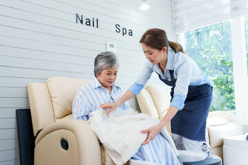 Obraz premium Asian female foot spa shop owner placing blanket on elderly woman in nail spa. Attentive, caring service in bright, cozy environment. client comfort and satisfaction in small business setting.
