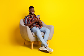 Full body portrait of nice young man sit chair use phone empty space wear shirt isolated on yellow color background