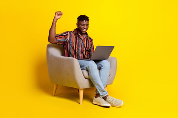 Full body portrait of nice young man sit chair laptop raise fist wear shirt isolated on yellow color background