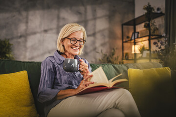 Fototapeta premium Mature woman sit in the living room read a book and drink coffee