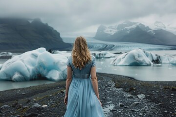 A woman in a blue dress stands on a rocky beach, looking out at the ocean