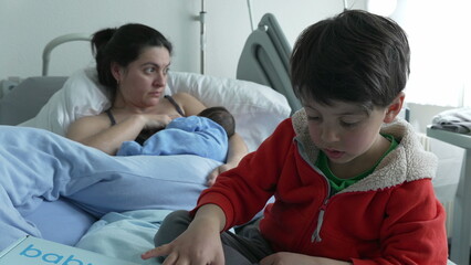 Mother breastfeeding newborn while older sibling in red jacket sits nearby on hospital bed....
