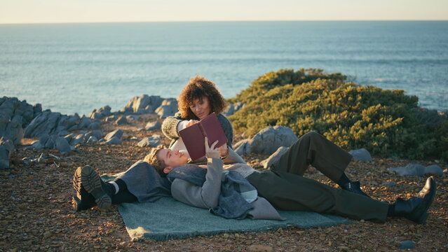 Relaxed couple lying picnic blanket at marine nature. Man reading book loud