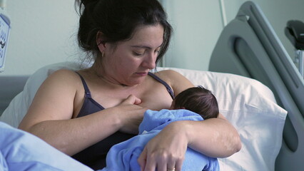 Mother breastfeeding newborn baby in a blue onesie, sitting in a hospital bed, showcasing a...