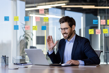 Smiling businessman in online meeting, waving hand at laptop screen. Professional setting in modern office with sticky notes on glass wall, productivity and communication concept