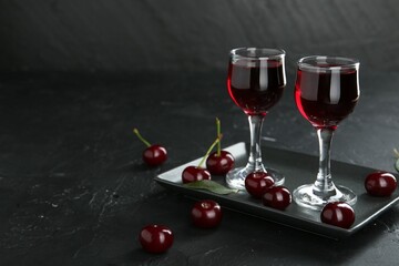 Delicious cherry liqueur in glasses and berries on black table. Space for text