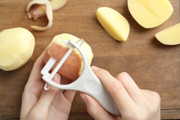 Woman peeling fresh potato with peeler at wooden table, top view