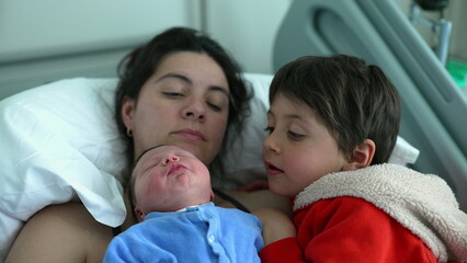 Older sibling tenderly kisses newborn baby while mother rests nearby, highlighting a loving family moment that emphasizes the bond between siblings and the nurturing presence of their mother