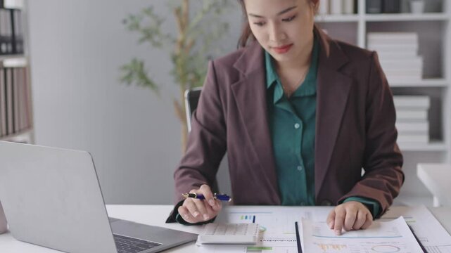 Businesswoman reviews financial graphs and calculates data at a tidy workspace with a laptop in a bright, modern office environment