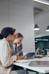 Two business women discuss online content and use phone and laptop while working in office