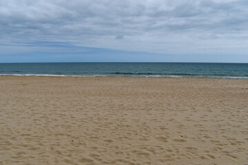 Wide shot of empty beach, sand on the shore, calm sea in the distance, cloudy sky