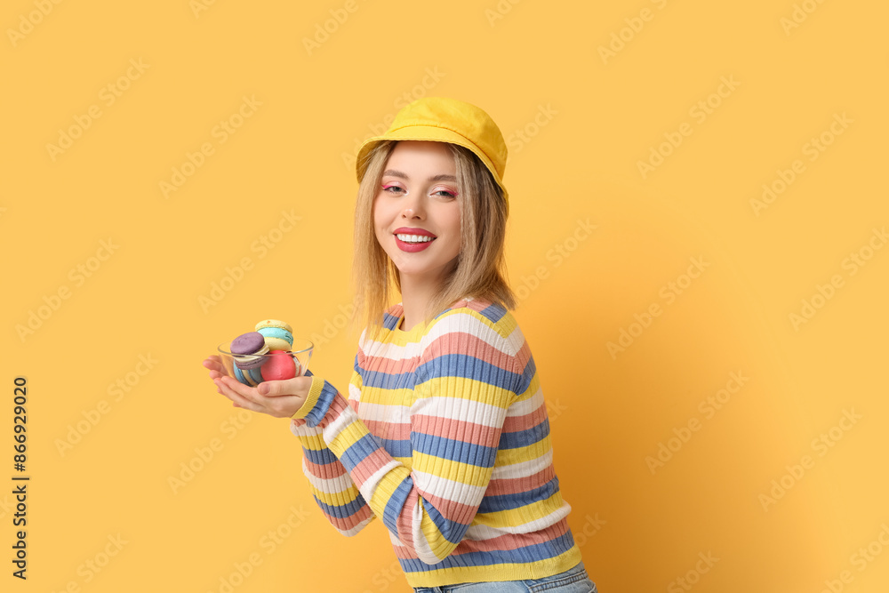 Poster Happy young woman holding glass bowl with sweet macaroons on yellow background