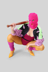 Beautiful young stylish woman in balaclava with baseball bat sitting on grey background