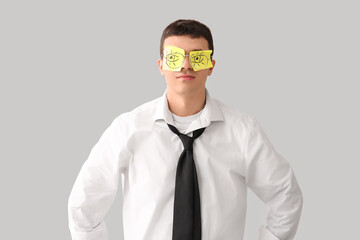 Tired young businessman with drawn eyes on sticky papers against light background