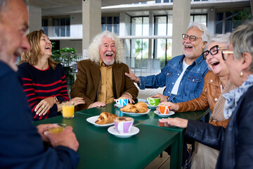 Group seniors Caucasian people laughing together sitting at cafeteria table. Old friends with gray hair having fun eating breakfast on terrace. Lifestyle of happy matures in the nursing home cafe