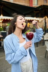 A woman is eating a fruit skewer while holding a glass of wine