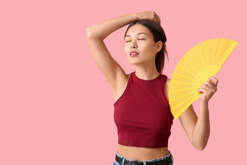 Young woman with hand fan suffering from heat on pink background